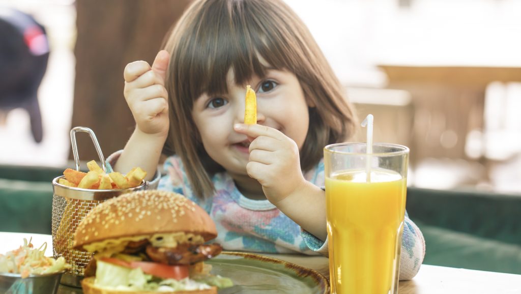 Little cute girl eating a fast food sandwich with fries and orange juice in a cafe. Fast food concept.