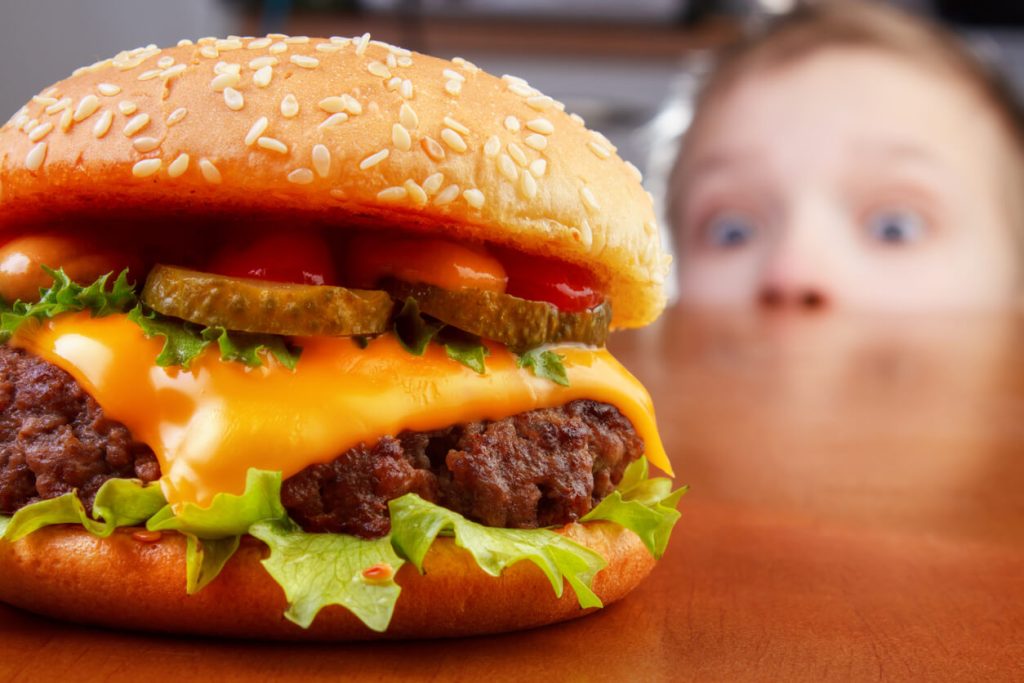 A little boy peeking greedily at a yummy hamburger on the table