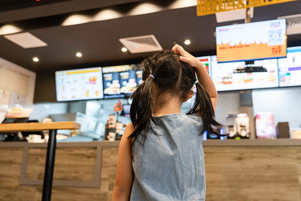 A little girl scratching her head in confusion at the fast food menu in front of her