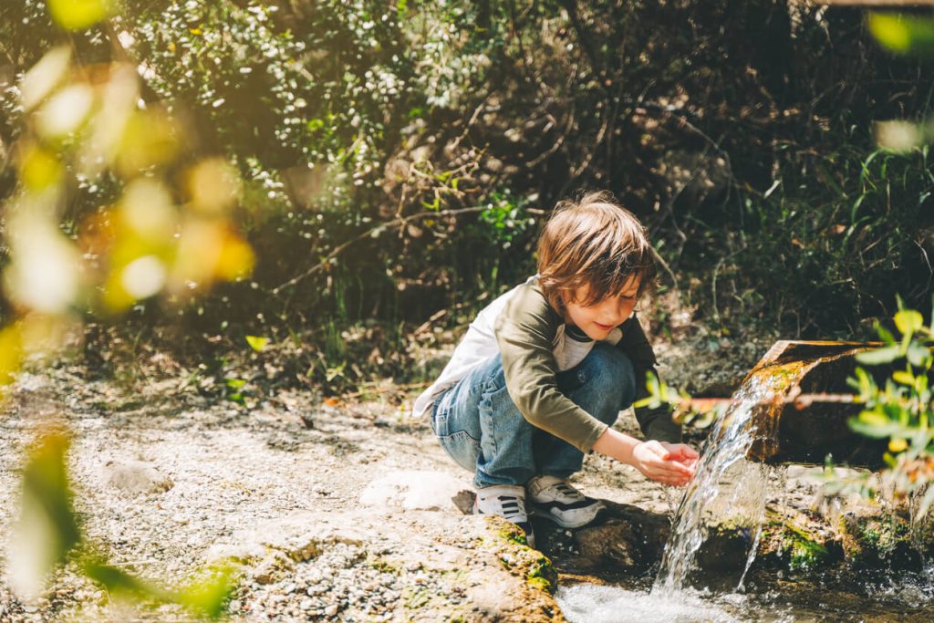 A young boy with his hands in a small water fall coming from a pipe in the forest.