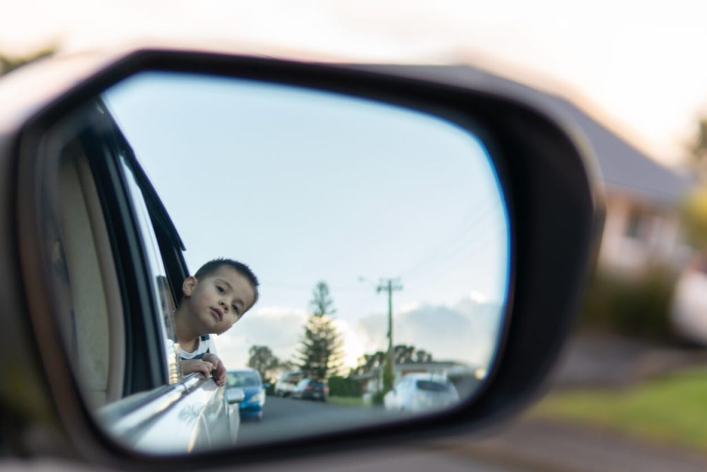 A little boy studying the side mirror of a car while he's in the back seat.